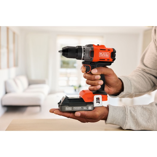A person holds a BLACK+DECKER cordless drill with an 18V battery, shown indoors with a blurred living room background.