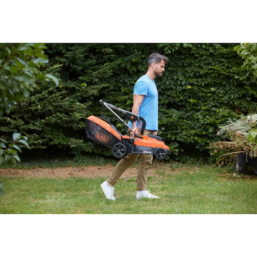 BLACKDECKER BCMW33184L2 lawn mower carried by person outdoors, orange and black body, garden setting.