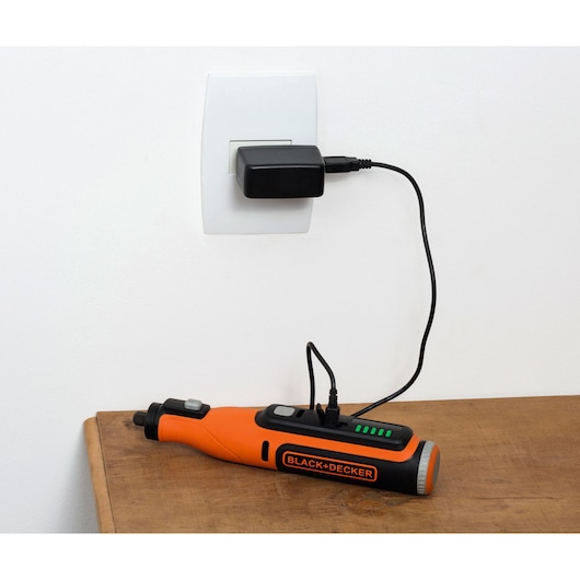 BLACK+DECKER BCRT8I cordless rotary tool charging on a wooden surface with adapter plugged into a wall socket.