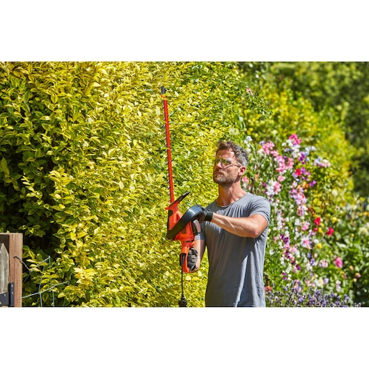 Photo of BLACKDECKER BEHTS551 hedge trimmer in use trimming a tall green hedge in a garden.