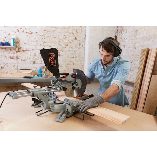 Person using a BLACK+DECKER BES710 sliding mitre saw to cut a wooden plank in a workshop. The person is wearing gloves and hearing protection.