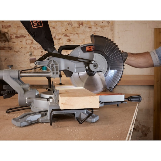 A BLACK+DECKER sliding compound miter saw is being used to cut a piece of wood on a workbench in a workshop environment. The saw blade is positioned over the wood, and a person wearing a glove is operating the tool.