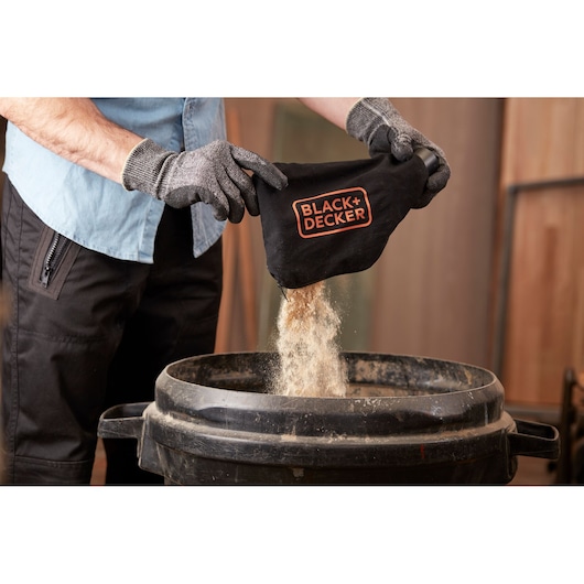 A person wearing gloves empties sawdust from a BLACK+DECKER dust bag into a large black bin.
