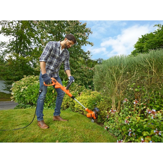 Photo of person using BLACK+DECKER BESTA528 orange electric grass trimmer in a garden, cable visible.