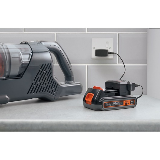 A BLACK+DECKER 18V lithium battery charger is placed on a countertop next to a cordless vacuum cleaner. The charger is plugged into a wall socket and is charging a battery. The scene is set against a tiled kitchen wall.