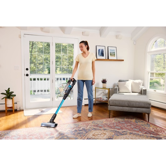 A person is vacuuming a living room with a BLACK+DECKER cordless stick vacuum cleaner. The room has a large window, a gray