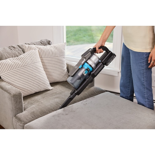A person cleaning a light grey upholstered chair with a BLACK+DECKER cordless stick vacuum cleaner. The vacuum has a