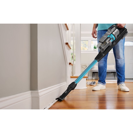 A person uses a BLACK+DECKER cordless stick vacuum cleaner to clean along the baseboard of a wooden floor near stairs in a