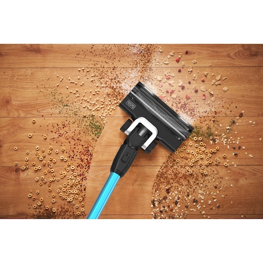 A BLACK+DECKER cordless stick vacuum cleaner with a blue handle is cleaning up a variety of scattered crumbs and dry debris