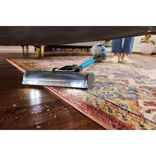 A cordless vacuum cleaner with LED lights is being used to clean under a sofa, showing its ability to reach under furniture