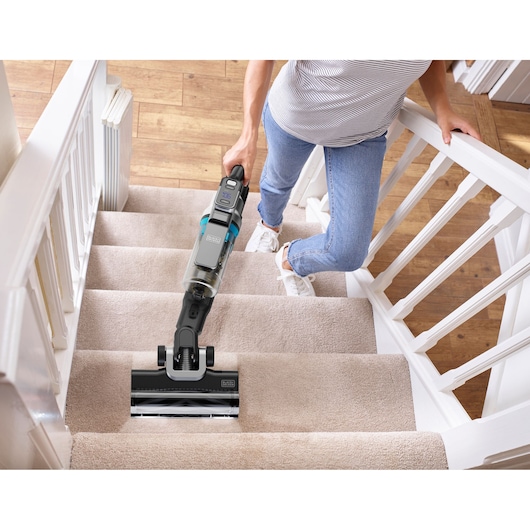 A person is cleaning carpeted stairs with a BLACK+DECKER cordless vacuum cleaner. The vacuum has a modern design and is