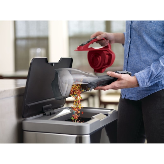 BLACKDECKER DVA315J handheld vacuum being emptied into a kitchen trash bin, colorful debris visible.