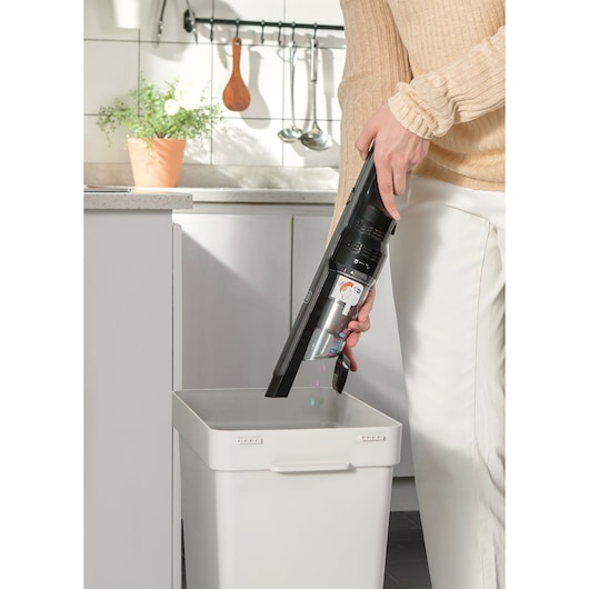 BLACKDECKER DVC320B21 handheld vacuum cleaner being emptied into a white trash bin in a kitchen.