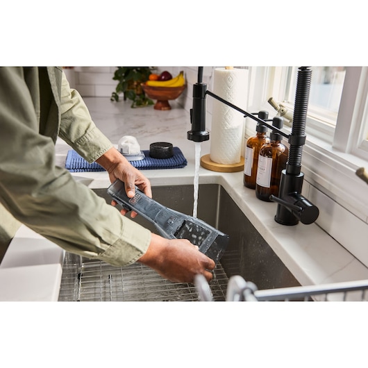 Person rinsing BLACKDECKER DVD320J vacuum dustbin under kitchen sink faucet.