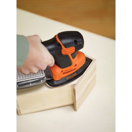 A person is using a BLACK+DECKER Mouse sander to sand the corner of a wooden box. The tool is orange and black, and it is labelled 'MOUSE.'