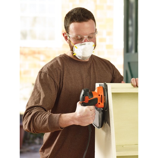 Person using an orange and black BLACK+DECKER electric sander to sand the side of a piece of wooden furniture. The sander is plugged in and the person is holding the furniture steady.