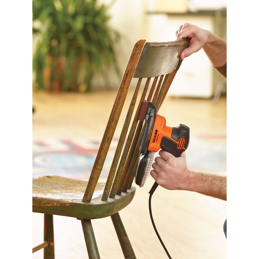 A person uses a BLACK+DECKER KA2000KA10 electric sander to sand the backrest of a wooden chair indoors.