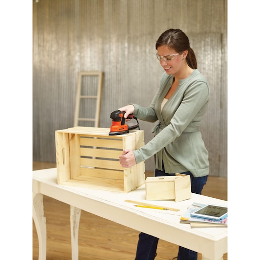 A person using a BLACK+DECKER KA2000KA10 electric sander to smooth a wooden crate on a white table. Additional wooden boxes, tools, and a tablet are also visible on the table.