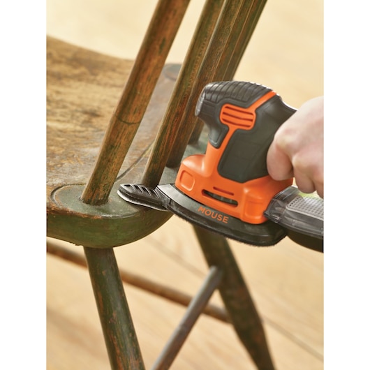 A close-up of a hand using a BLACK+DECKER KA2500K mouse sander to sand the wood of a chair, focusing on detailed and hard-to-reach areas.
