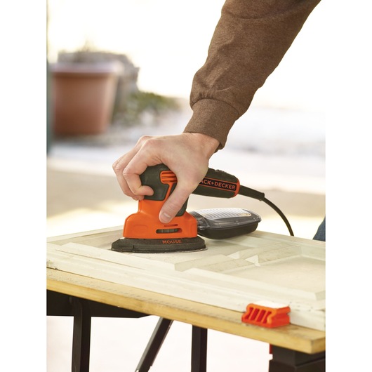 A person uses an orange and black BLACK+DECKER KA2500K electric sander to smooth the surface of a wooden panel on a workbench.