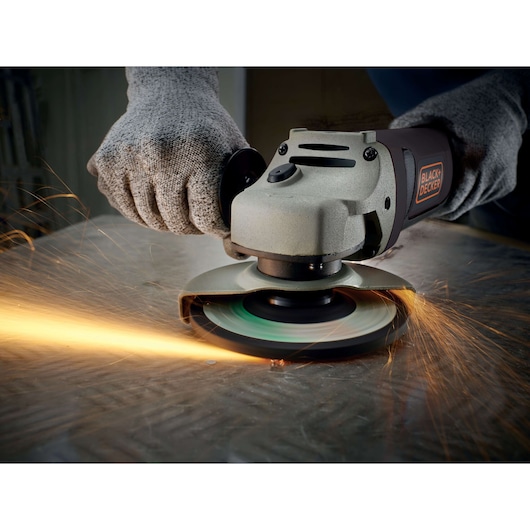 Close-up of a BLACK+DECKER angle grinder being used to cut metal, with sparks flying. The user is wearing protective gloves.