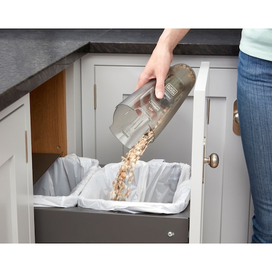 BLACK+DECKER NVC115W handheld vacuum canister being emptied into a kitchen trash bin.