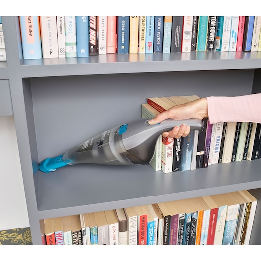 BLACK+DECKER NVC215WA handheld vacuum with blue nozzle cleaning a gray bookshelf, books visible in background.