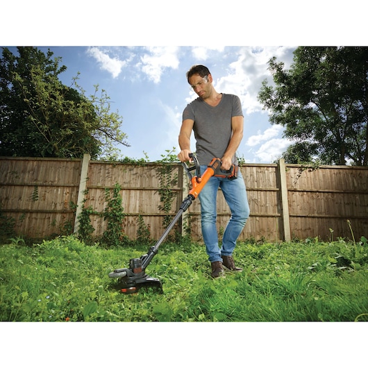Photo of person using BLACKDECKER STC1840EPC cordless grass trimmer in a garden with overgrown grass.