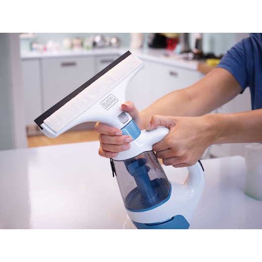 Photo of BLACK+DECKER WW100 window vacuum cleaner held by hands in a modern kitchen setting.